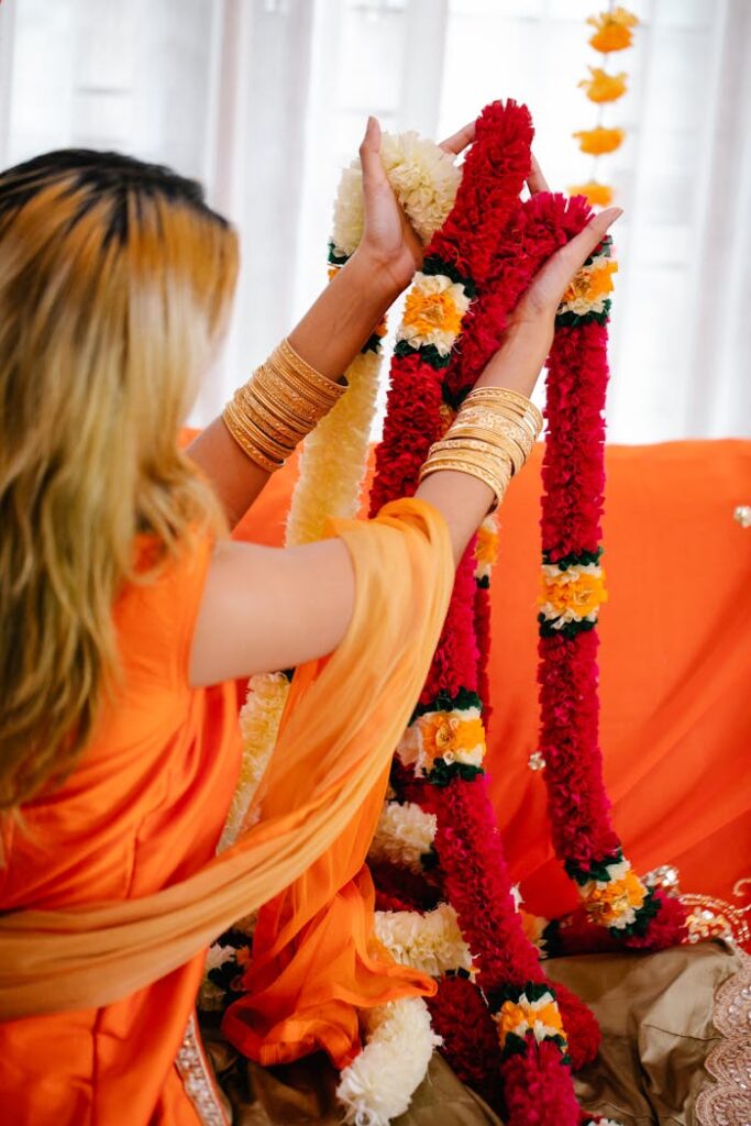 pexels photo 8887094 Back view of a woman in orange holding vibrant floral garlands in a festive setting.
